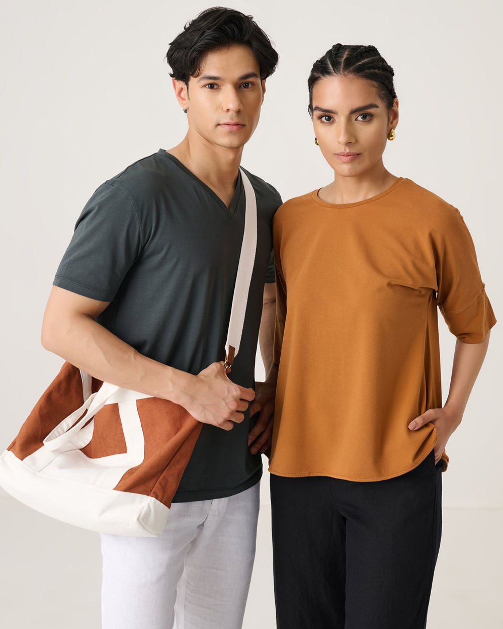 Man and woman standing side by side, man holding a bag, wearing a dark gray shirt and light gray pants, woman wearing a brown shirt and black pants.